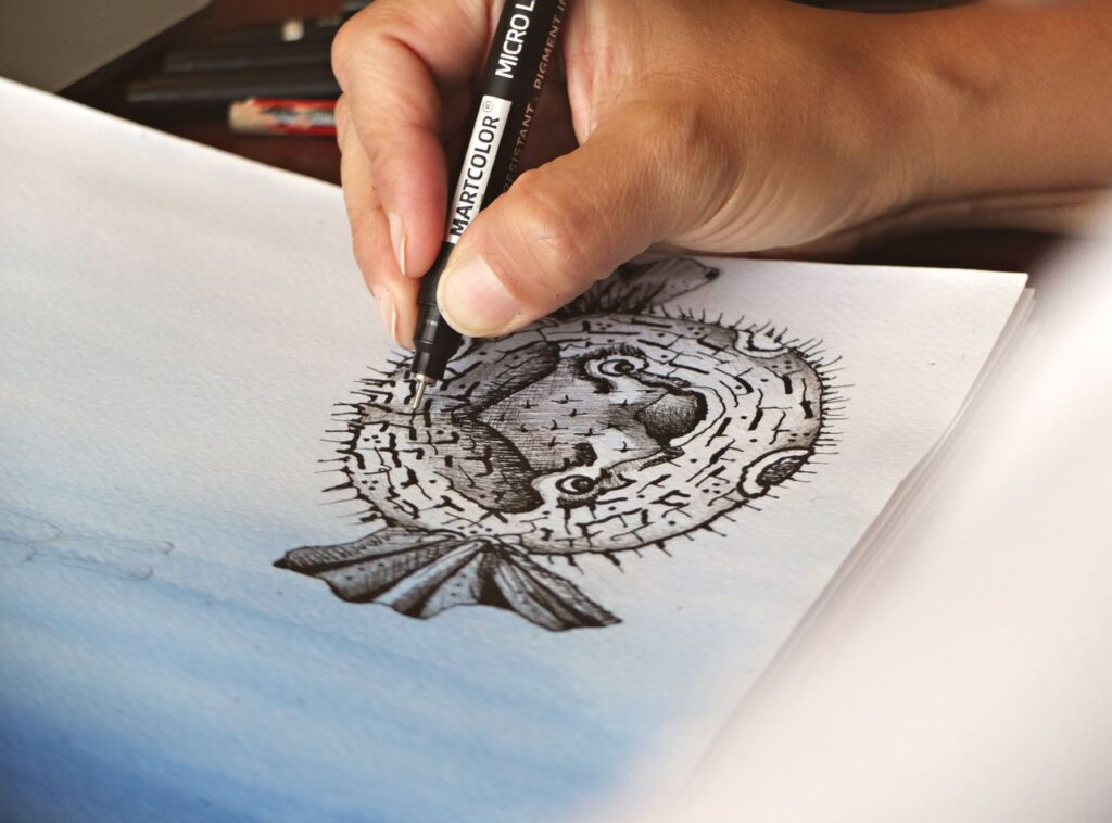 Close-up of a hand drawing a pufferfish with a fineliner pen on paper.