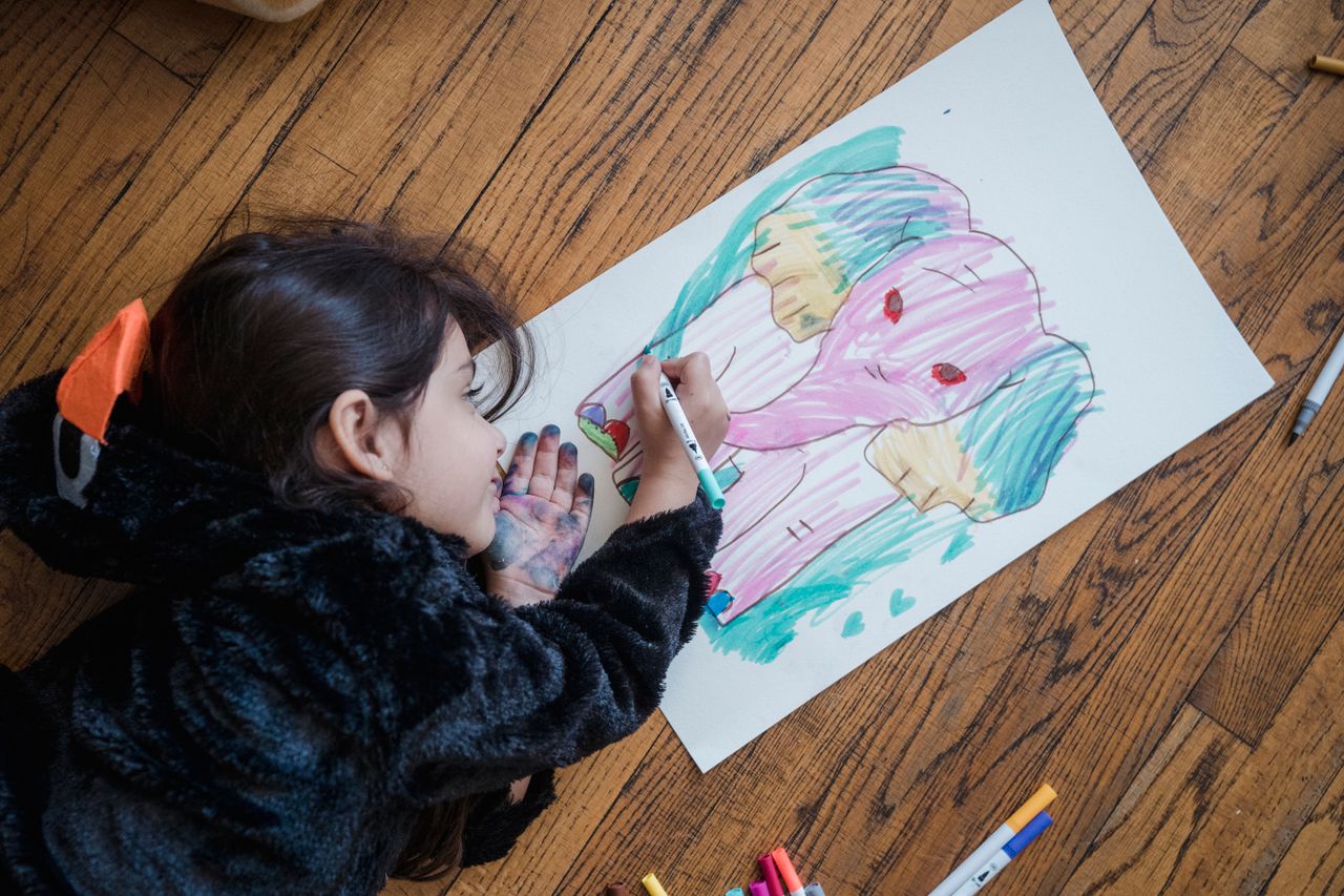 Young girl drawing an elephant with markers on wooden floor, showcasing creativity and innocence.